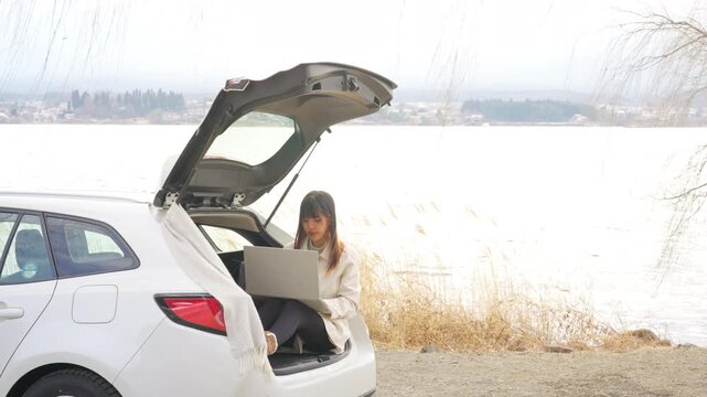 Asian woman using laptop while sitting in open car trunk by lakeside in Japan. Female traveler remote working during road trip, workation, digital nomad lifestyle and winter travel holiday vacation