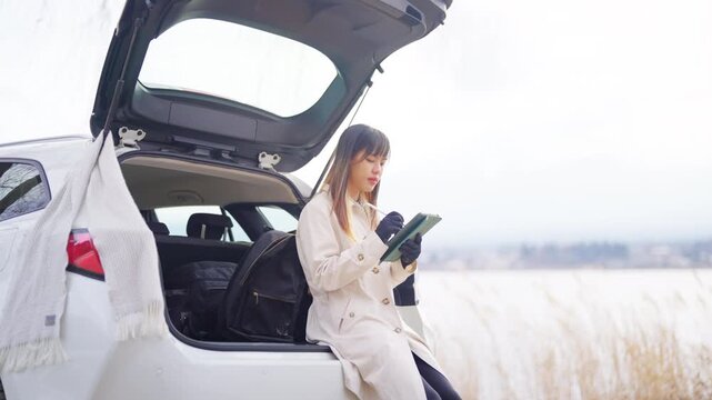 Asian woman using digital tablet while sitting in car trunk by lakeside in Japan. Female traveler remote working during road trip, workation, digital nomad lifestyle and winter travel holiday vacation