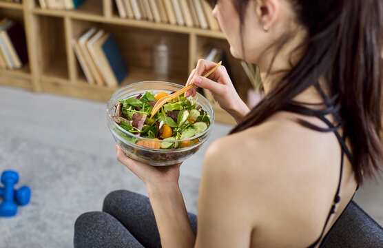 Woman eating healthy salad at home. A fit female in sportswear holds a bowl of fresh vegetables and greens, sitting a cozy room, illustrating fitness, diet, nutrition, and mindful weight control.