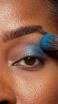 Close up of professional makeup artist applying blue eyeshadow on dark skin model eyelid.