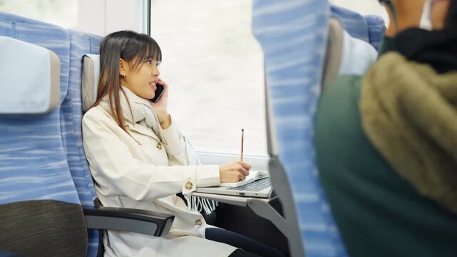 Asian businesswoman talking on smartphone and remote working on laptop while business travel by train. Female professional conference call mobile office during railway transportation commute workation