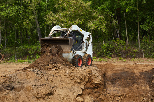 Compact skid steer loader pushing and dumping soil on a construction site near a forest