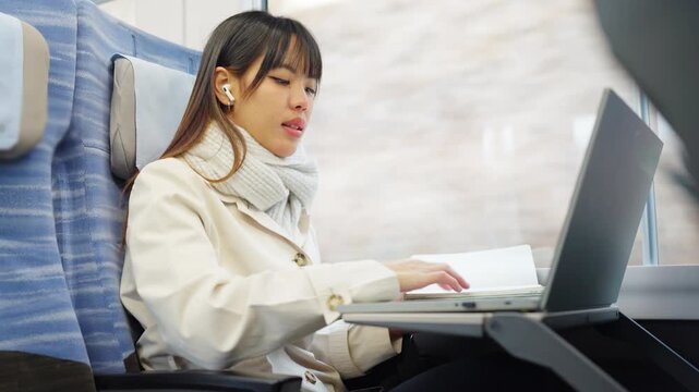 Asian businesswoman using wireless earbuds conference call and remote working on laptop while business travel by train. Female professional hands free communication, workation and mobile office