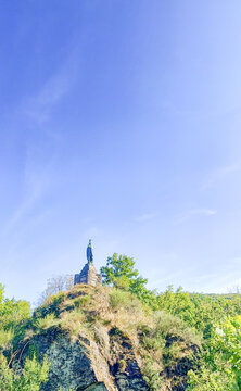 Esch-sur-Sure, Wiltz, Grand-Duche de Luxembourg, September 07, 2025, Church spire rises behind hilltop statue with lush green foreground winding path and distant village rooftops under clear blue sky