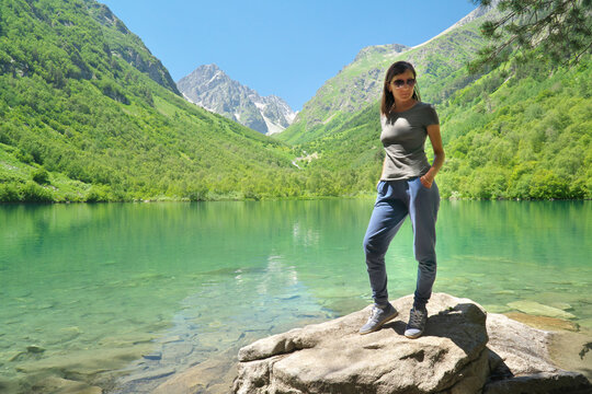 Young woman in sportswear stands on a rock by a turquoise mountain lake with green slopes under a blue sky. Alpine landscape suits travel