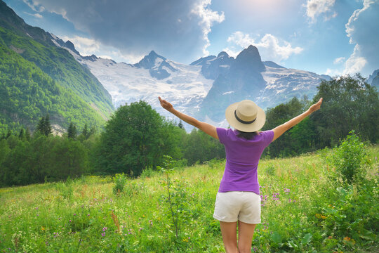 Young female tourist in a straw hat raising her arms toward majestic snowy mountains from a green meadow. Inspiring eco travel and freedom