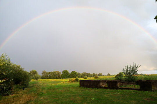 A full colorful rainbow arc spans the sky above green fields, trees, and an old concrete foundation after a rainstorm. This panoramic rural landscape conveys tranquility, hope, and natural splendor.