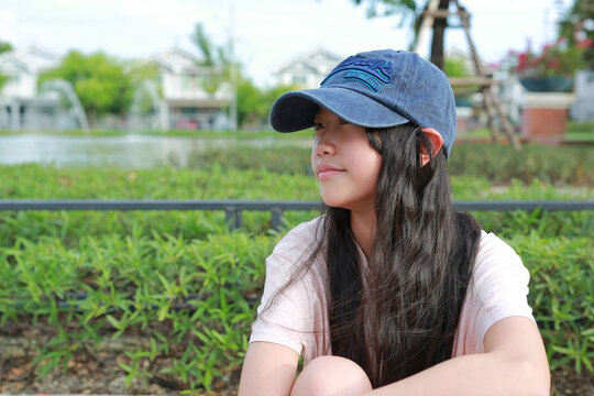 A young Asian girl wearing a blue baseball cap sitting outdoors in a park, looking away thoughtfully while relaxing.