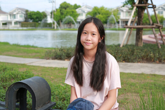 A young Asian girl is sitting in a park by a lake and smiling.