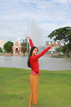 A young Asian girl wearing a red cardigan sweater and yellow pants standing outdoors in a park with raised arms and posing happily in front of a fountain and lake.