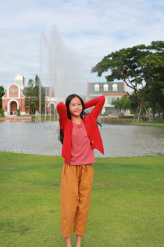 Cute Asian girl in red cardigan standing on grass with her hands behind her head, relaxing in a park with a fountain and European-style building in background.