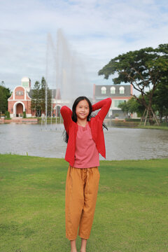 A young Asian girl standing on green lawn in a park, placing her hands behind her head and posing in front of a lake with a fountain.