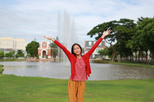 Happy young Asian girl smiling and raising her hands in air, expressing joy and freedom, standing on grassy field in front of a park, with trees, a pond, fountains, and building background.