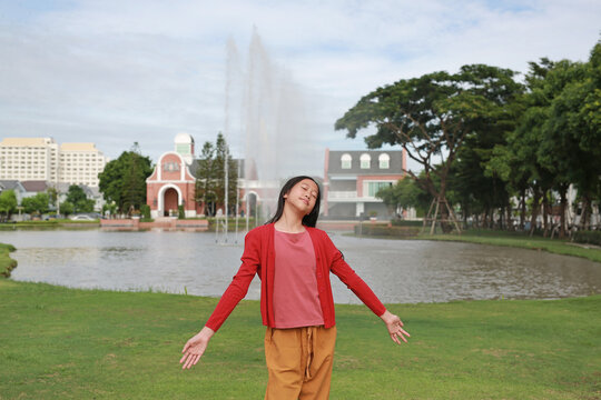 An Asian girl stands with her arms outstretched and eyes closed, taking a breath of fresh air in front of a fountain and buildings in a park.