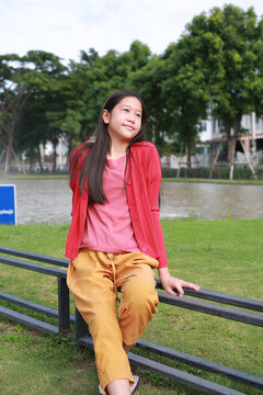 Joyful young Asian girl sitting comfortably on metal fence in an outdoor park setting, smiling warmly while looking up towards against grassy field, trees, and a body of water.