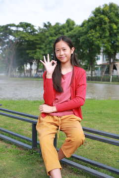 Portrait of young Asian girl sitting on bench in a park, smiling and making an 'okay' hand sign.