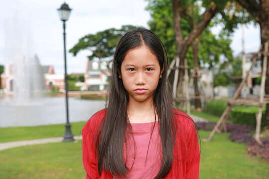 A young Asian girl with long dark hair, She is in an outdoor park setting with green grass, trees, and a blurred background featuring a fountain.