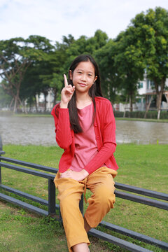 A young Asian girl in red cardigan sits on bench in a park, pointing her index finger upward with a gentle smile.