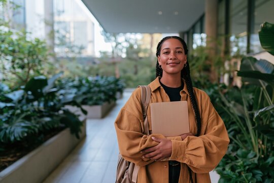 Student with a deep skin tone holding a laptop in a campus concourse, wearing a textured mustard overshirt for university admissions and student life campaigns