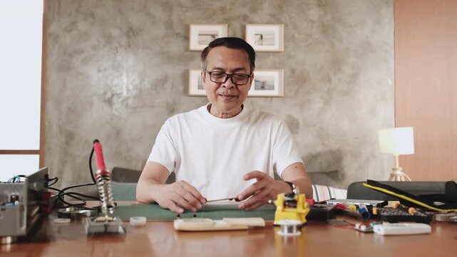 Mature Asian man showing pride in circuit board after successful soldering job, multimeter check, cheerful job achievement, home electronics appliance repair handyman, DIY fix tools, hobby workshop.