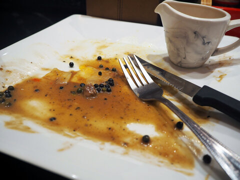 A dirty plate after a meal, with leftover sauce and peppercorns, and used cutlery. A small jug sits on the side.