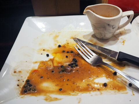 Remnants of a delicious meal on a white plate, with a small pitcher of sauce beside a fork and knife. The plate is speckled with black peppercorns and coated in a savory brown sauce.