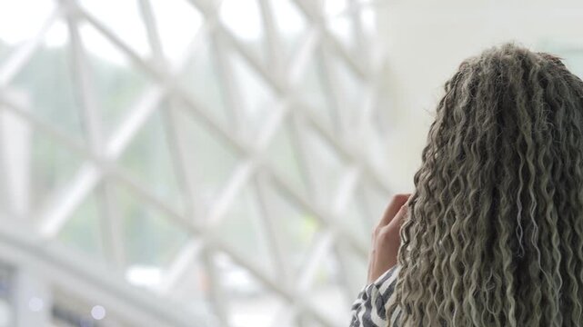 Woman with curly hair looks out a window. Her hand rests near her face. She stands in a modern, bright space. The glass wall lets in soft daylight. This clip suits travel, waiting