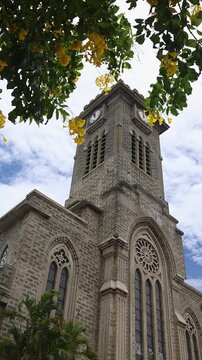 Low angle view of the iconic stone tower and facade of the historic Nha Trang Cathedral framed by blooming yellow flowers and green foliage against a partly cloudy sky. Travel and tourism, Vietnam.