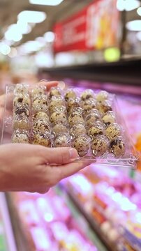 Close-up of a woman hands holding and choosing a transparent plastic carton package of fresh speckled quail eggs from display shelves in a grocery store or supermarket. Retail shopping, food.