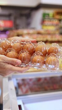 Close-up of a woman hands holding and choosing a transparent plastic carton package of fresh brown chicken eggs from display shelves in a grocery store or supermarket. Retail shopping, food.