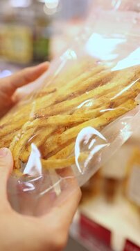 Close-up of a woman hands holding and choosing a vacuum-sealed plastic package of dried fish strips or snacks from display shelves in a grocery store or supermarket. Retail shopping, food.