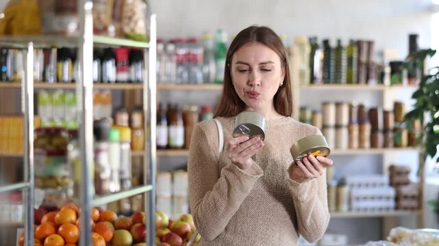Young woman holding two tin cans of preserves in hands, choosing and shop around pate for sandwiches. She carefully studies information for consumer, thinks about buying
