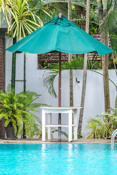 Green umbrella shading a white poolside table beside a resort swimming pool surrounded by lush tropical plants