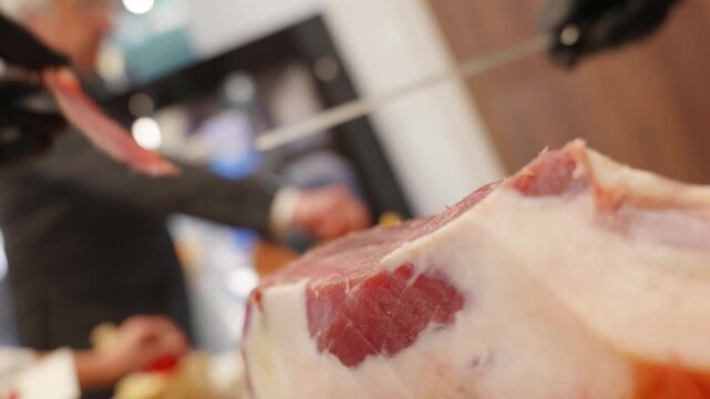 Extreme close-up of a chef slicing thin pieces of prosciutto crudo by hand with a long knife while wearing black protective gloves.