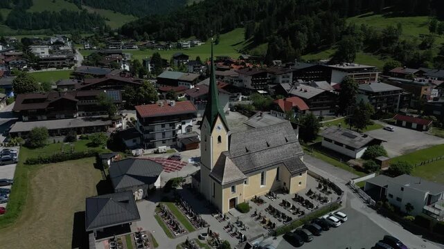 Reveal Shot on a clear, sunny day of a traditional Austrian mountain village. The central focus is a historic Roman Catholic church