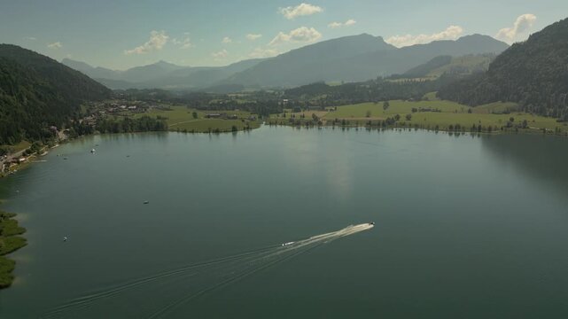 A high-angle drone photograph capturing a vast, calm mountain lake nestled in a valley. In the lower right foreground, a motorboat speeds across the water,