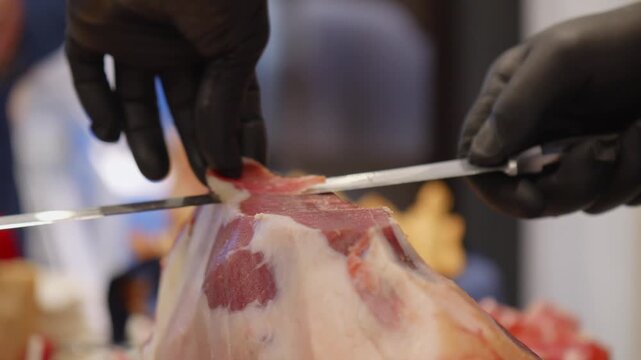 Close-up of a chef carefully cutting thin slices from a whole prosciutto crudo using a long carving knife and black gloves.