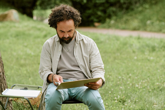 Middle aged Caucasian man drawing on clipboard during outdoor art master class in park. Image represents creative hobbies and is suitable for leisure or education marketing