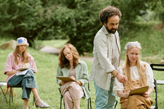 Middle aged Caucasian male art teacher guiding diverse young adult women drawing on clipboards during outdoor master class, promoting creative education and recreational workshops