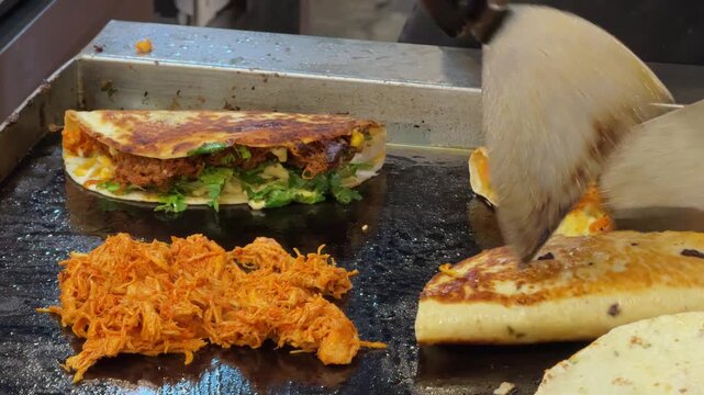 Close-up footage of a food vendor preparing multiple fresh tacos and wraps on a hot flat-top griddle. Tortillas are filled with meat, cheese, vegetables, herbs and sauces during busy street food prepa