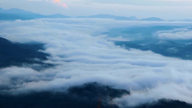 High-angle panning shot sweeping across a vast sea of thick white fog and clouds covering deep mountain valleys during early morning twilight.