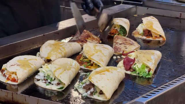 Close-up footage of a food vendor preparing multiple fresh tacos and wraps on a hot flat-top griddle. Tortillas are filled with meat, cheese, vegetables, herbs and sauces during busy street food prepa