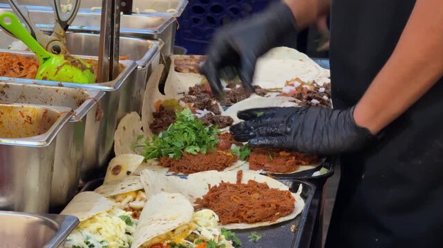 Close-up footage of a food vendor preparing multiple fresh tacos and wraps on a hot flat-top griddle. Tortillas are filled with meat, cheese, vegetables, herbs and sauces during busy street food prepa