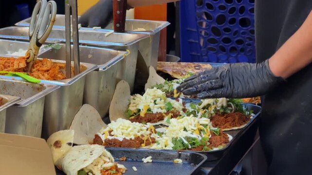 Close-up footage of a food vendor preparing multiple fresh tacos and wraps on a hot flat-top griddle. Tortillas are filled with meat, cheese, vegetables, herbs and sauces during busy street food prepa