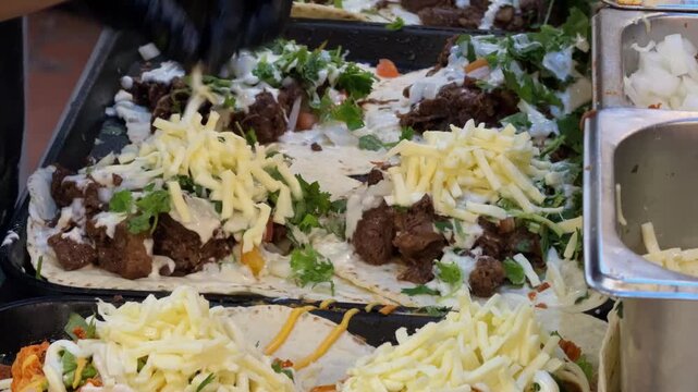 Close-up footage of a food vendor preparing multiple fresh tacos and wraps on a hot flat-top griddle. Tortillas are filled with meat, cheese, vegetables, herbs and sauces during busy street food prepa