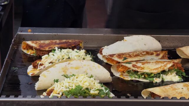 Close-up footage of a food vendor preparing multiple fresh tacos and wraps on a hot flat-top griddle. Tortillas are filled with meat, cheese, vegetables, herbs and sauces during busy street food prepa