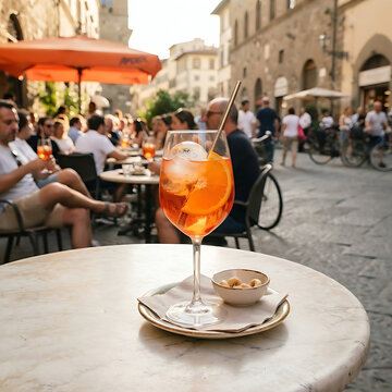A bright orange Aperol Spritz with a large ice sphere, garnished with a fresh orange slice, served in a stemmed wine glass. The scene is a bustling European cafe terrace in the late afternoon sun.