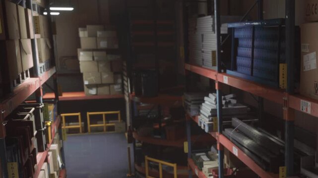 Workers move around the warehouse filled with shelves stacked with boxes and equipment. The layout shows pathways and organized storage space. Natural light comes from above.