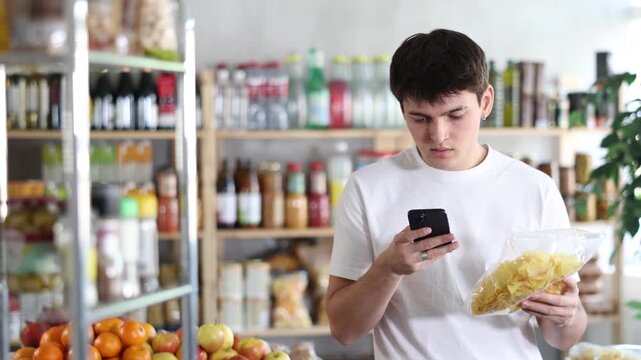 Young guy scanning bag of crispy chips with smartphone at grocery store, using mobile payment app for cashless transaction. Concept of modern seamless shopping and digital convenience. High quality 4k