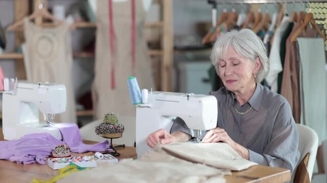 Older dressmaker works at a sewing machine against the background of racks with fabrics. Experienced clothing designer at work in a sewing workshop . High quality 4k footage